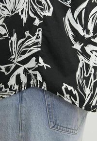 Tela negra con un diseño floral abstracto en blanco, con un dobladillo curvado. Denim azul claro visible por debajo, con un bolsillo trasero.