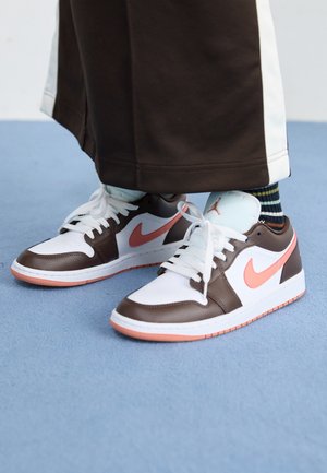 Baskets Nike blanches et marron avec des virgules roses, portées avec un pantalon foncé et des chaussettes rayées sur un sol bleu.