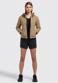 Giacca cappuccio marrone chiaro con chiusura a zip, due tasche frontali e una texture liscia. Indossata sopra una maglia nera e shorts di denim scuro.