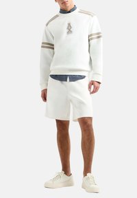 Weißer Pullover mit braunen Streifen, Logodetail, kombiniert mit weißen Shorts. Gestylt mit einem blauen karierten Hemd darunter und weißen Sneakern.