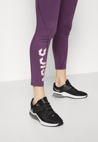 Zapatillas deportivas negras con parte superior de malla, diseño de cordones y amortiguación de aire visible. Llevadas con mallas moradas que tienen texto de logo blanco.