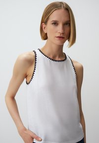 Witte mouwloze top met een ronde halslijn, verfraaid met zwarte zigzagstiksels langs de randen. Glad materiaal, relaxt model.