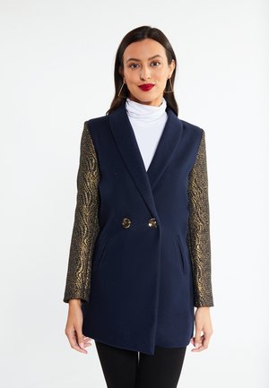 Nainen pukeutuneena laivastonsiniseen kaksiriviseen bleiseriin, jossa on kultakuvioidut hihat, valkoinen poolopaita ja geometriset kultaiset korvakorut, seisoo valkoista taustaa vasten.