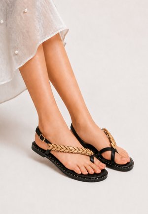 Mujer con sandalias negras y doradas con tiras en forma de hoja y una falda blanca translúcida con perlas, de pie sobre un suelo claro.