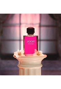 Rosa glasparfymflaska med en svart kapsyl märkt "JOOP! HOMME," placerad på en vit dekorativ pelare med vita blommor.