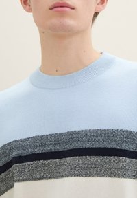 Hellblauer Strickpullover mit Rundhalsausschnitt, der graue und marineblaue horizontale Streifen über der Brust aufweist. Glatte Textur, keine auffälligen Akzente.