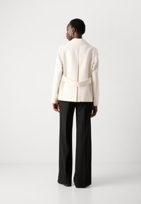 Lichtbeige blazer met een gestructureerd ontwerp, voorzien van een rugriem en een split. Gecombineerd met hooggerokte zwarte broeken van een gladde stof.