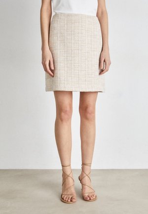 Kvinde iført nederdel i beige tekstureret stof og knælængde, hvid top og nude-farvede sandaler med stropper, stående mod en ensfarvet lys baggrund.