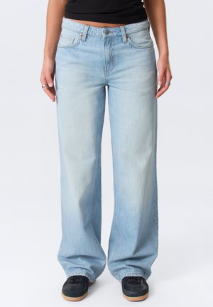 Osoba ubrana w jasnoniebieskie szerokie jeansy z wysokim stanem, czarne trampki z gumowymi spodami oraz czarny krótki top, stojąca na jednolitym tle.