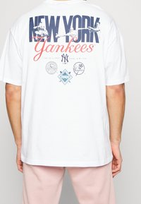 Biała koszulka bawełniana z grafiką 'NEW YORK Yankees' w granatowym i czerwonym kolorze, z wieloma logo na plecach w różnych odcieniach kolorystycznych.