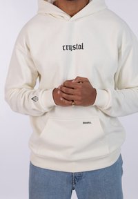 Sudadera crema con texto negro "crystal" y "dreams." en la parte frontal. Cuenta con un bolsillo tipo canguro y un logo de diamante en la manga. Tejido suave y texturizado.