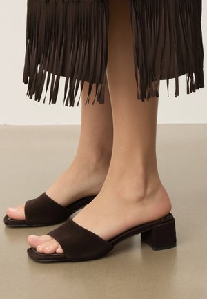 Sötétbarna, nyitott orrú, blokksarkú papucsot viselő lábak, barna rojtos szoknyával párosítva, sima padlón.