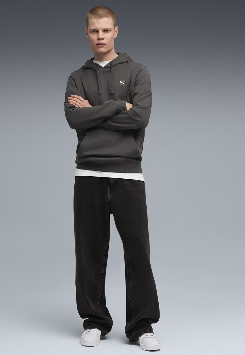 Felpa grigia con cappuccio e un piccolo logo, abbinata a jeans neri larghi e sneakers bianche, su uno sfondo grigio sfumato.