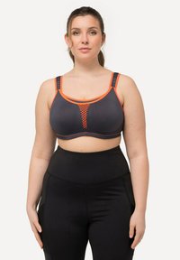 Sujetador deportivo gris con detalles en naranja, que presenta un inserto de malla y tirantes anchos. Combinado con leggings negros de cintura alta. Textura suave.