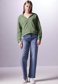 Felpa verde a zip con texture, vestibilità rilassata, indossata con jeans dritti azzurri e sneakers bianche. Design semplice, senza pattern visibili.