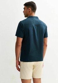 Camisa de manga corta en un color verde oscuro con un patrón texturizado, cuellos abotonados y un acento en forma de bucle en la parte posterior, combinada con pantalones cortos de color claro.