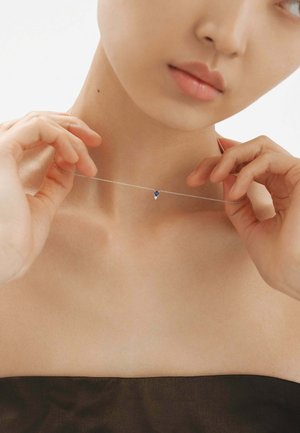 Collar de cadena de plata delicada con un pequeño acento de gema azul, reposando sobre la clavícula, con manos ajustándolo suavemente.