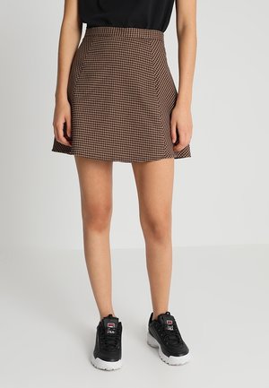 Femme portant une mini-jupe pied-de-poule marron, un haut noir et des baskets noires FILA, debout devant un fond clair uni.