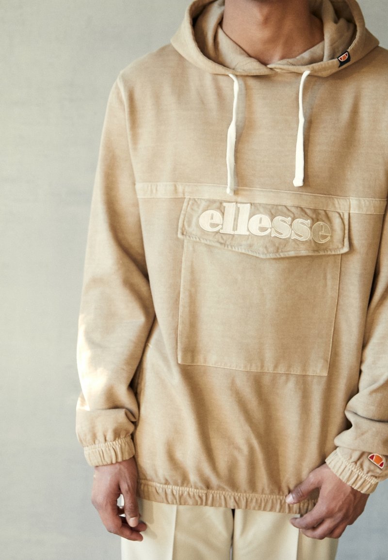 Sweat à capuche beige avec des poignets élastiques, poche avant et logo "ellesse" brodé. Texture douce avec un subtil éclat.