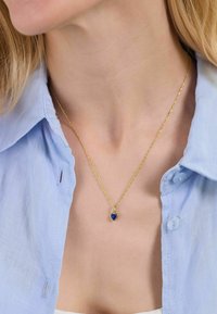 Collar de oro con una cadena retorcida y un pequeño colgante en forma de corazón azul, llevado sobre una camiseta de color azul claro.