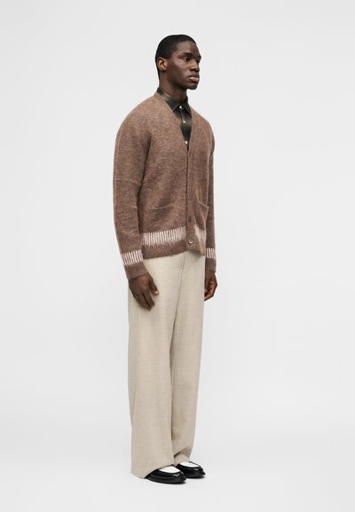 Άνδρας που στέκεται, φοράει καφέ χνουδωτή ζακέτα πάνω από σκούρο πουκάμισο, μπεζ παντελόνι με φαρδιά πόδια και μαύρα-λευκά μοκασίνια, σε λευκό φόντο.