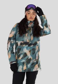 Osoba v různobarevném maskáčovém kabátu, černých rukavicích, černých kalhotách, béžové čepici a lyžařských brýlích s purpurovými skly.