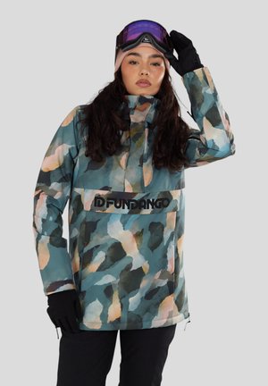 Person in einer mehrfarbigen Tarnjacke, schwarzen Handschuhen, schwarzen Hosen, einer beige Mütze und Skibrille mit lila Gläsern.