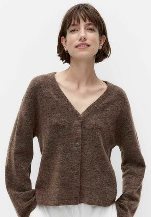 Vrouw met kort bruin haar draagt een bruine, zachte, geknoopte cardigan met V-hals en lange mouwen tegen een effen lichte achtergrond.