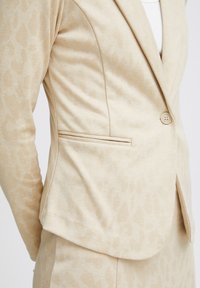 Béžový blazer s jemným leopardím vzorom, štruktúrovaný dizajn, zapínanie na jeden gombík a bočné vrecká. Mäkka, textúrovaná tkanina.