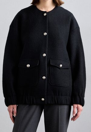 Frau trägt eine schwarze Wolljacke mit silbernen Knöpfen und zwei vorderen Klappentaschen über grauen Hosen, steht vor einem schlichten Hintergrund.