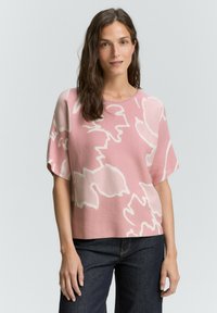 Růžový pletený top s bílým květinovým vzorem, krátkými rukávy, širokým výstřihem a volným střihem. V kombinaci s tmavými džínovými kalhotami.