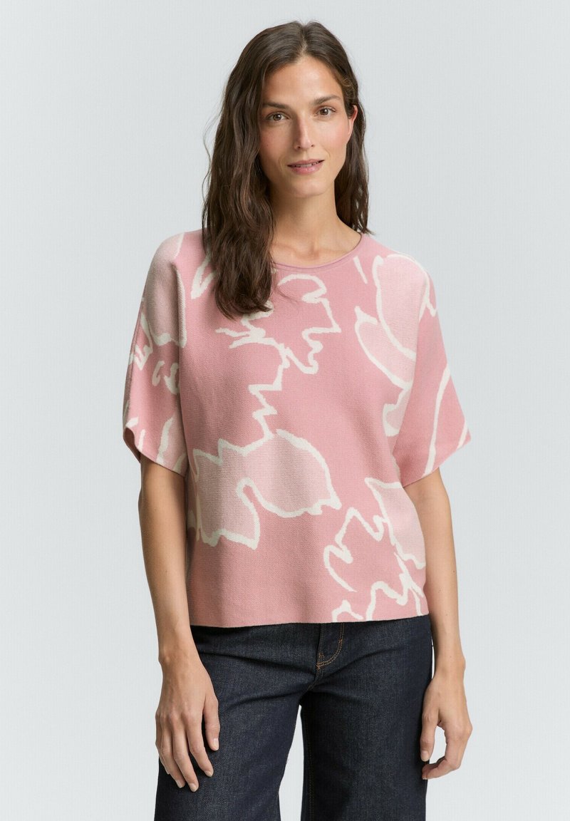 Růžový pletený top s bílým květinovým vzorem, krátkými rukávy, širokým výstřihem a volným střihem. V kombinaci s tmavými džínovými kalhotami.