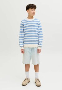 Garçon portant un pull rayé bleu et blanc, un short en denim clair, des chaussettes blanches et des chaussures beige, debout et regardant vers la droite sur un fond uni.