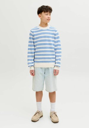 Zēns zilā un baltā svītrainā džemperī, gaišās džinsa šortos, baltās zeķēs un bēšās kurpēs, stāvot un skatoties pa labi uz vienkārša fona.