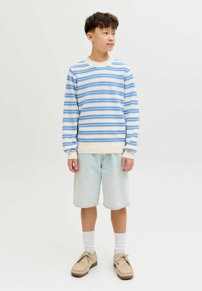 Garçon portant un pull rayé bleu et blanc, un short en denim clair, des chaussettes blanches et des chaussures beige, debout et regardant vers la droite sur un fond uni.