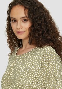 Oliwkowa bluzka z białym wzorem w kropki, okrągły dekolt i krótkie rękawy; miękki materiał z widocznymi detalami szwów.