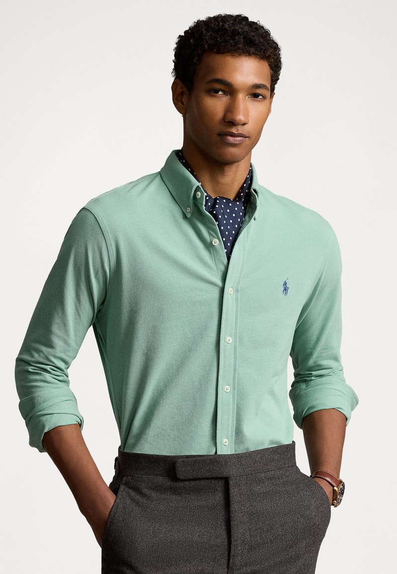 Camisa de manga longa verde claro com colarinho abotoado, apresentando um pequeno logótipo azul no peito. Usada por cima de uma camisa azul marinha com bolinhas.