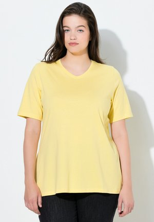 Junge Frau mit braunen Haaren, die ein hellgelbes T-Shirt mit V-Ausschnitt und kurzen Ärmeln sowie eine dunkle Hose trägt und vor einer weißen Wand steht.