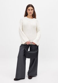 Weißer gerippter Pullover mit weitem Ärmel, kombiniert mit dunkelgrauen, weiten Hosen. Dunkle schwarze Lederhandtasche mit abgerundetem Griff.