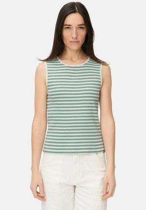 Frau trägt ärmelloses, grün-weiß gestreiftes Oberteil und weiße Hose, steht vor hellem einfarbigem Hintergrund.