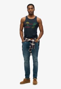 Marineblauw tanktop met een grafisch ontwerp van palmbomen en zonsondergang, gecombineerd met blauwe jeans en een geruit shirt om de taille geknoopt. Bruine schoenen.