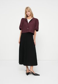 Blonde woman in a textured burgundy short-sleeve jacket, white top, black pleated midi skirt, and black low-heeled slingback shoes standing.