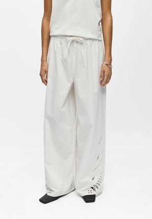 Femme portant un pantalon large blanc ample avec des détails découpés brodés et une taille à cordon, associé à des chaussures noires à bout carré.