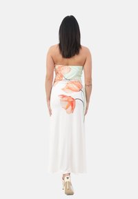 Femme aux longs cheveux noirs portant une robe blanche sans bretelles avec un imprimé floral orange, s'éloignant en chaussures à talons blancs sur un fond uni.