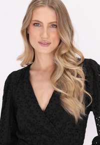 Schwarzes Wickelkleid mit floraler Lochmusterung, V-Ausschnitt und Puffärmeln. Das Model hat lange, gewellte blonde Haare und ein neutrales Makeup.
