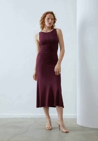 Vestido sem mangas em burgundy com detalhes em franzido, ajustado na parte superior e a alargar na bainha. Usado com sapatos de salto dourados com tiras.