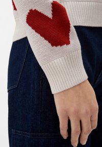 Stickad tröja med ett rött hjärtmönster på en ljus bakgrund, med ribbade ärmslut och en texturerad väv, ihop med mörka jeansbyxor.