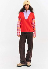 Czerwona i fioletowa bluza z suwakiem przy szyi, rękawy w pomarańczowo-fioletową kratę, zestawiona z brązowymi spodniami sztruksowymi i beżowymi botkami na sznurowadła.