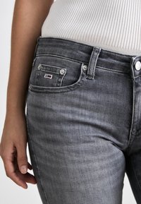 Harmaat farkut, joissa on istuva muotoilu. Niissä on pieni etutasku, metallinapit ja logo-etiketti punaisena, valkoisena ja sinisenä taskussa.