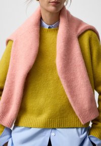 Geel wollen sweater met een geribbelde onderrand, gecombineerd met een zachte roze wollen sjaal die om de schouders is gedrapeerd over een lichtblauw shirt.